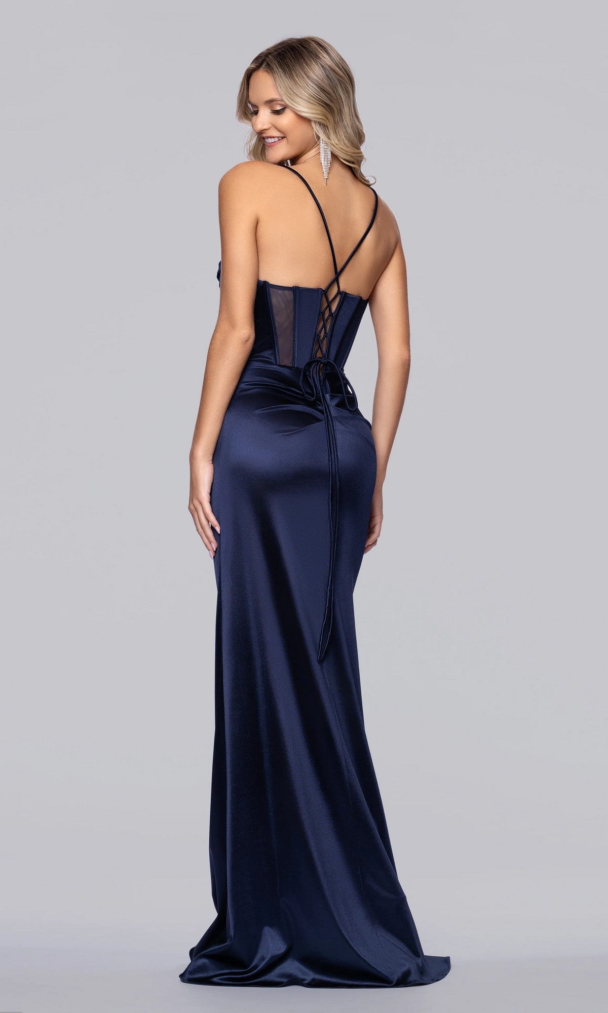 A woman wearing the Blondie Nites 6014BN long navy satin prom dress with spaghetti straps, lace-up back, and floor-length skirt stands looking over her shoulder against a plain background.